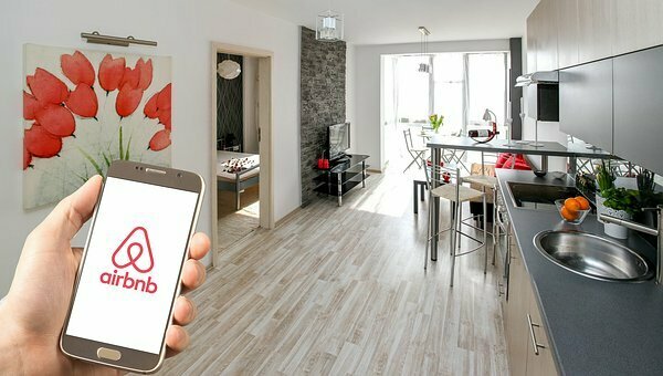 Смартфон в руке с приложением AIRBNB на фоне кухни-гостиной