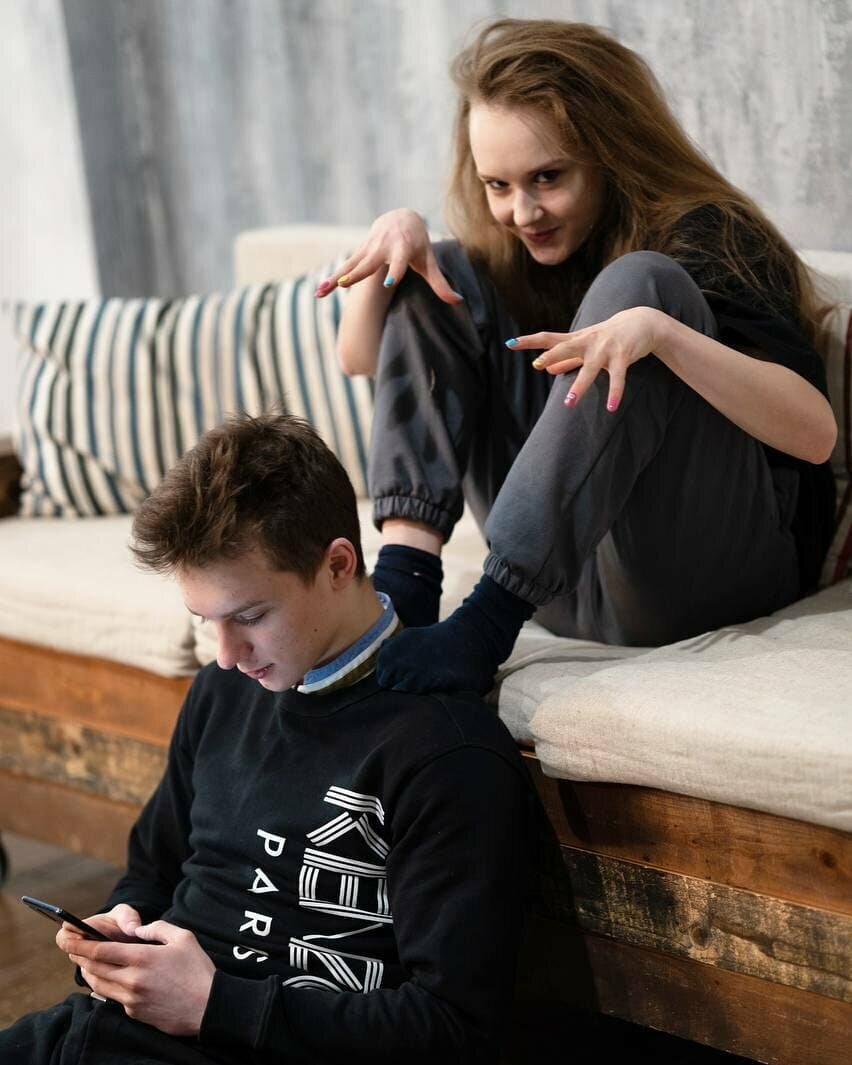 Илья уткнулся в телефон, сидит на полу. Ангелина сидит на диване, поместила ноги на плечи и делает вид, что пугает партнёра - https://www.instagram.com/angelina_k21